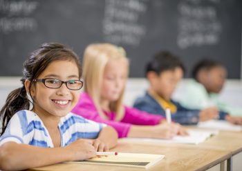 A multi-ethnic group of elementary age children are sitting together in class at their desks. One girl is smiling and looking at the camera.